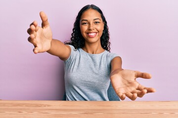 Wall Mural - Young african american girl wearing casual clothes sitting on the table looking at the camera smiling with open arms for hug. cheerful expression embracing happiness.