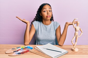 Wall Mural - Young african american girl artist sitting at studio table clueless and confused with open arms, no idea and doubtful face.