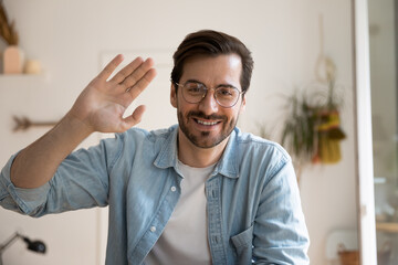 Smiling millennial male in glasses posing before digital webcam waving hand chatting with friend using pc. Headshot portrait of active young man video blogger looking at camera broadcasting from home