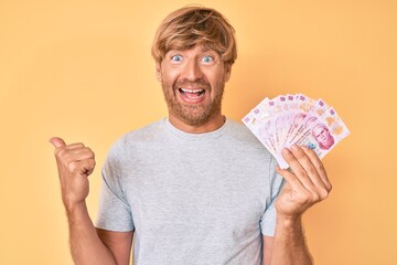 Sticker - Young blond man holding mexican pesos pointing thumb up to the side smiling happy with open mouth