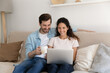 © fizkes - Calm relaxed millennial couple spend free evening weekend on cozy sofa drink tea coffee watch movie using laptop. Smiling young husband wife engaged in choosing goods at web store chat with friends