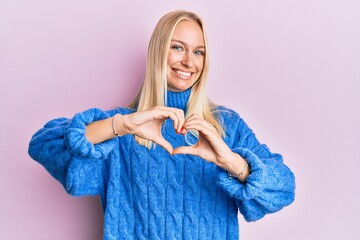 Poster - Young blonde girl wearing wool winter sweater smiling in love doing heart symbol shape with hands. romantic concept.