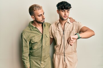 Poster - Homosexual gay couple standing together wearing casual jumpsuit checking the time on wrist watch, relaxed and confident