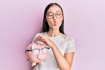 Poster - Young chinese woman holding piggy bank with glasses making fish face with mouth and squinting eyes, crazy and comical.