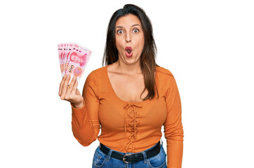 Sticker - Beautiful hispanic woman holding 100 yuan chinese banknotes scared and amazed with open mouth for surprise, disbelief face