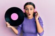 © Krakenimages.com - Young hispanic woman using headphones holding vinyl disc afraid and shocked with surprise and amazed expression, fear and excited face.