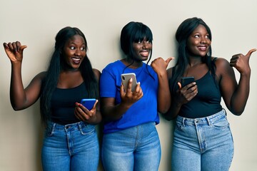 Three young african american friends using smartphone smiling with happy face looking and pointing to the side with thumb up.