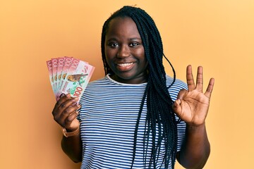 Sticker - Young black woman with braids holding 100 new zealand dollars banknote doing ok sign with fingers, smiling friendly gesturing excellent symbol