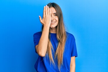 Young hispanic woman wearing casual clothes covering one eye with hand, confident smile on face and surprise emotion.