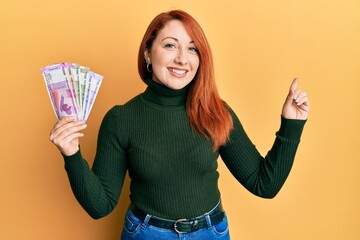 Sticker - Beautiful redhead woman holding indian rupee banknotes smiling happy pointing with hand and finger to the side