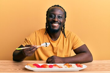 Poster - Handsome young black man eating sushi sitting on the table looking positive and happy standing and smiling with a confident smile showing teeth