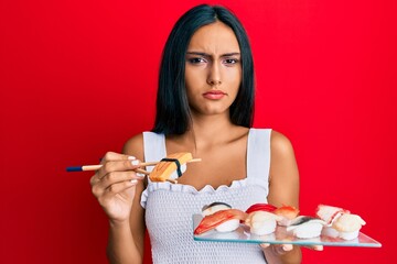 Poster - Young brunette woman eating omelet sushi using chopsticks clueless and confused expression. doubt concept.