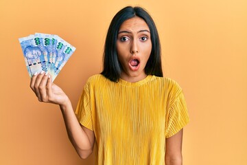 Poster - Young brunette woman holding south african 100 rand banknotes scared and amazed with open mouth for surprise, disbelief face