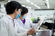 © bung - Young junior scientist wear hygiene mask and lab coat in the modern laboratory looking at colleague. Woman using tablet and show the result . Research and education concept.