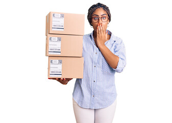 Canvas Print - Young african american woman with braids holding delivery packages covering mouth with hand, shocked and afraid for mistake. surprised expression