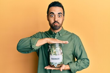 Canvas Print - Young hispanic man holding jar with savings puffing cheeks with funny face. mouth inflated with air, catching air.