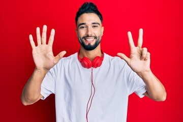 Poster - Young man with beard listening to music using headphones showing and pointing up with fingers number eight while smiling confident and happy.