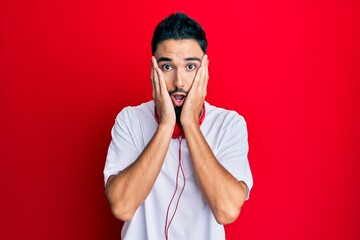 Canvas Print - Young man with beard listening to music using headphones afraid and shocked, surprise and amazed expression with hands on face