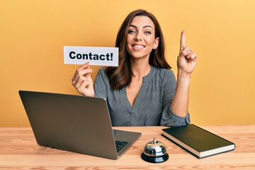 Wall Mural - Young brunette woman working at hotel reception holding contact paper smiling with an idea or question pointing finger with happy face, number one