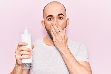 Wall Mural - Young handsome bald man holding bottle of cream lotion covering mouth with hand, shocked and afraid for mistake. surprised expression