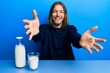 © Krakenimages.com - Handsome caucasian man with long hair holding glass of milk looking at the camera smiling with open arms for hug. cheerful expression embracing happiness.