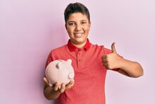 Teenager Hispanic Boy Holding Piggy Bank Smiling Happy And Positive, Thumb Up Doing Excellent And Approval Sign