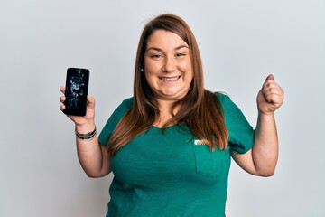 Beautiful brunette plus size woman holding broken smartphone showing cracked screen screaming proud, celebrating victory and success very excited with raised arm