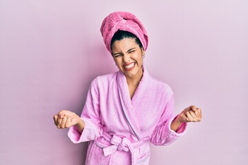 Young hispanic woman wearing shower towel cap and bathrobe very happy and excited doing winner gesture with arms raised, smiling and screaming for success. celebration concept.