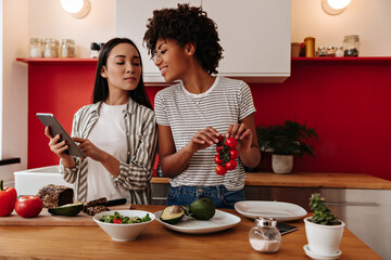 African girl with smile looks into tablet and holds tomatoes. Women posing in kitchen