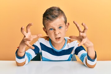 Adorable caucasian kid wearing casual clothes sitting on the table shouting frustrated with rage, hands trying to strangle, yelling mad