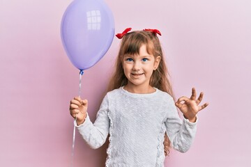 Little caucasian girl kid holding purple balloon doing ok sign with fingers, smiling friendly gesturing excellent symbol