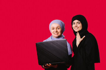 Two muslim female friends standing over red background with a laptop in hands. Young woman in with a covered heads looking to the camera and smiling 