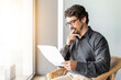 © Joao - Man wearing glasses on a chair holding paper document. Successful male portrait thinking and reading contract files at home office by the window