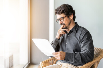 Wall Mural - Man wearing glasses on a chair holding paper document. Successful male portrait thinking and reading contract files at home office by the window