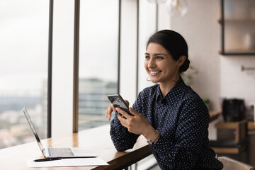 Canvas Print - Happy young indian lady work on laptop online at modern flat office look at large picture window dream holding mobile phone. Pretty smiling hindu woman excited with good news received in phone message