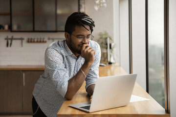 Wall Mural - Smiling black guy freelancer stand at kitchen desk read good email message on laptop screen think on advantages proposed by customer. Young afro man hipster chat with friends online from home using pc