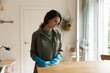 © fizkes - Young Caucasian woman housewife in rubber gloves clean kitchen countertop wooden surfaces. Millennial female housekeeper or wife do daily house routine, wipe dust from table furniture at home.