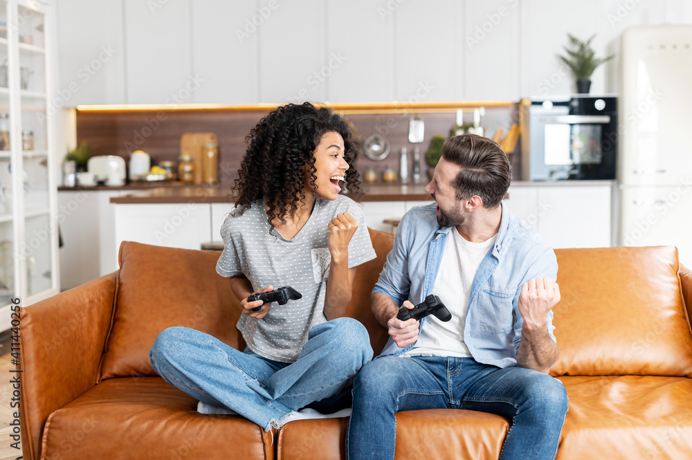 Excited multiracial couple enjoys playing video game, an african woman and a caucasian man hold wireless joysticks look at each other ready to fight. Interracial couple spends leisure time at home - obrazy, fototapety, plakaty 