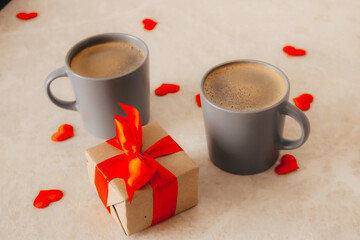 two gray coffee cups sit on the table, next to a gift box with a red bow and scattered red confetti hearts. The concept of St. Valentine's Day