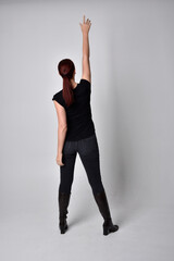 Canvas Print - Full length portrait of woman with red hair in a ponytail, wearing black t-shirt and denim pants. Standing pose, hands reaching out with back to the camera the camera, against a  studio background.