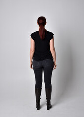 Wall Mural - Simple full length portrait of woman with red hair in a ponytail, wearing casual black tshirt and jeans. Standing pose with back to the camera the camera, against a  studio background.