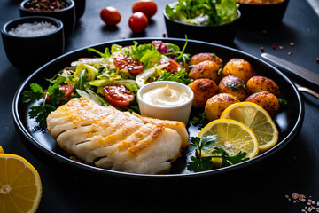 Fish dish - fried cod fillet with potatoes and vegetable salad on black wooden table
