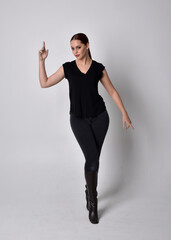 Poster - Simple full length portrait of woman with red hair in a ponytail, wearing casual black tshirt and jeans. Standing pose front on with hand reaching gestures, against a  studio background.