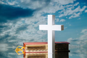 Holy bible and bright white cross against blue clouded sky. It's standing on a reflective surface.