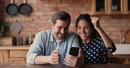 Wall Mural - Happy couple sit at table in kitchen read received sms on smartphone device, got great commercial special offer, profit price and discount. Good news, on-line casino winners, moment of victory concept