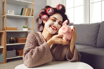 Happy funny young woman with beauty face mask on, in hair rollers and bathrobe, hugging pink piggy bank, smiling and daydreaming. Concept of loving money, making plans, and family budget management