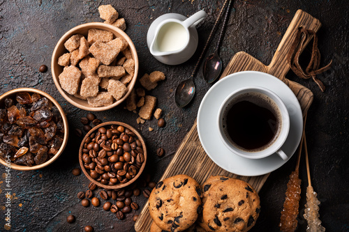 Cup of coffee with chocolate drop cookies, bowls of brown cane lump sugar, crystal sugar sticks, milk and coffee beans © viktoriya89