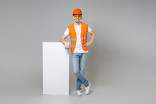 Full Length Young Employee Man In Orange Vest Protective Hardhat Lean On Blank Sign Board Place For Text Akimbot Isolated On Grey Background. Instruments For Renovation Apartment Room. Repair Concept.