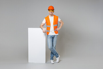 Full length young employee man in orange vest protective hardhat lean on blank sign board place for text akimbot isolated on grey background. Instruments for renovation apartment room. Repair concept.