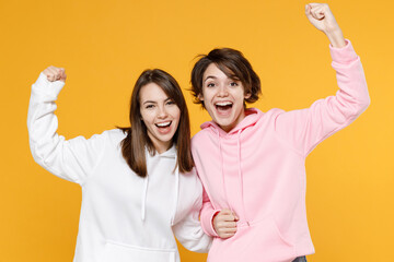 Cheerful happy two young women friends 20s wearing casual white pink hoodies doing winner gesture celebrating clenching fists say yes isolated on bright yellow color wall background studio portrait.